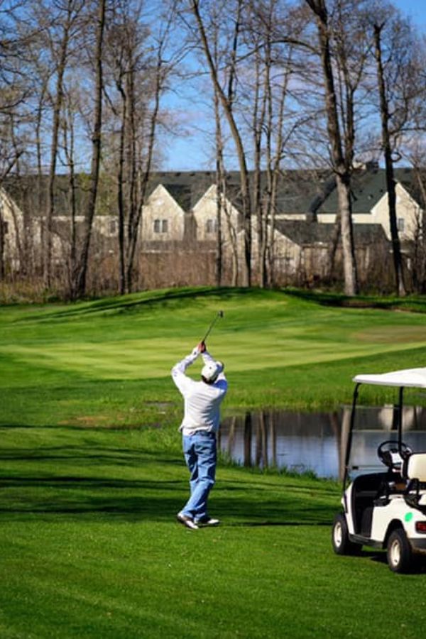A golfer swings beside a pond on a green, with a golf cart nearby.