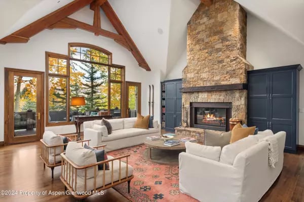 Living room with exposed timber beams, a tall stone fireplace, and large windows overlooking trees.