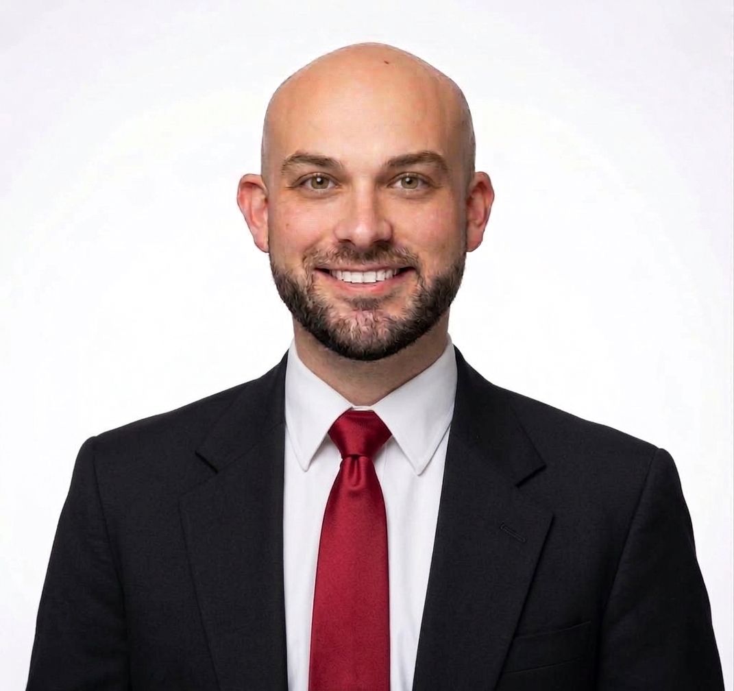 Mark Vaughn smiling in a black suit, white shirt, and red tie.
