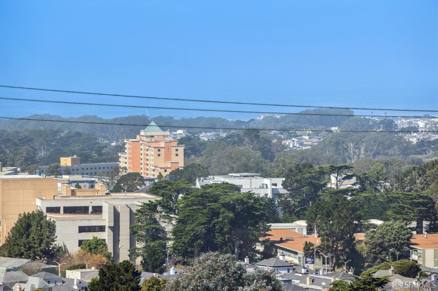 City buildings and trees with wooded hills in the distance beneath a clear blue sky.