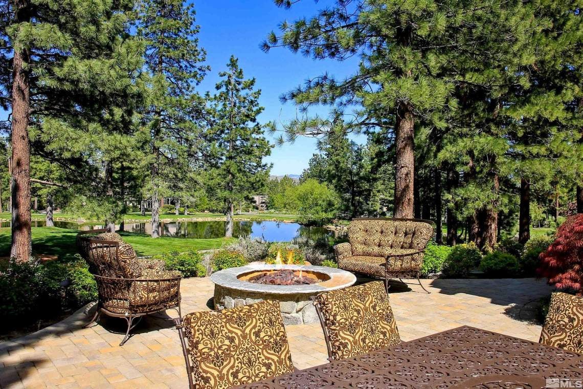 Stone patio with patterned seating and a fire pit overlooking a pond among pine trees.