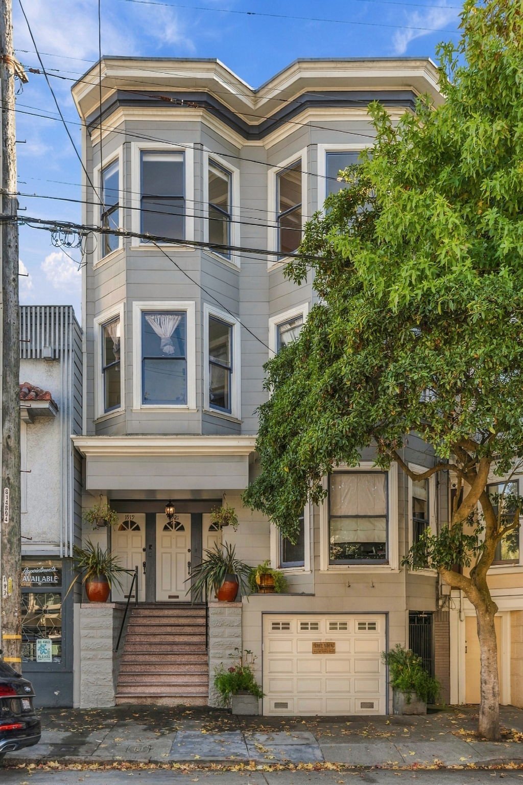 Gray multi-story residence with bay windows, front steps, garage, and a leafy tree along the sidewalk.
