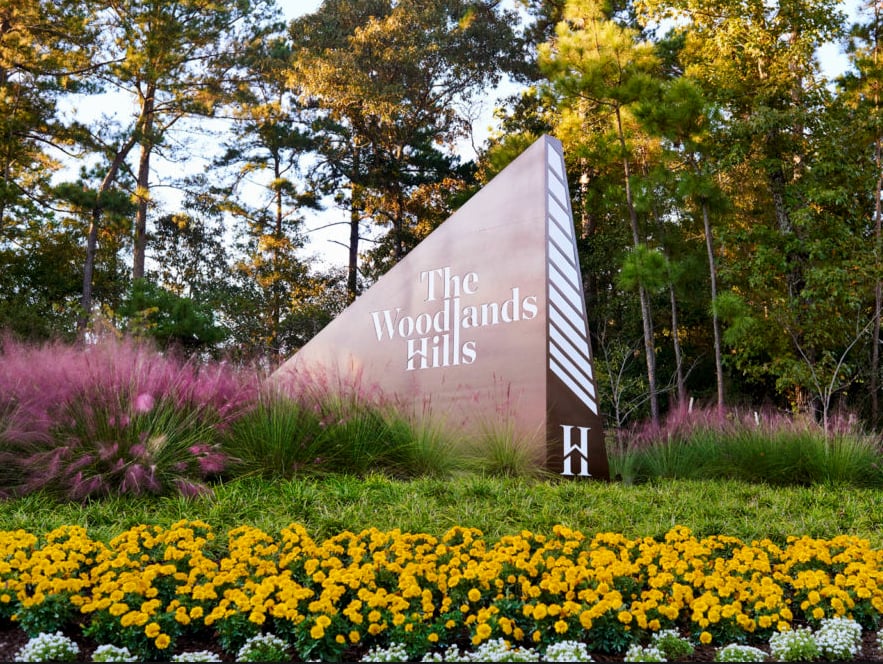 The Woodlands Hills entrance sign among trees and colorful flower beds.