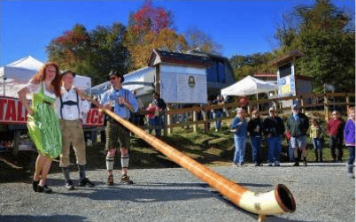 Festivalgoers hold a long wooden horn at an outdoor gathering beneath autumn-colored trees.
