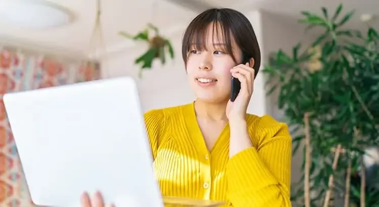 A smiling woman wearing a yellow shirt, holding a tablet or clipboard, and talking on the phone. The background is a well-lit indoor setting with plants, indicating a positive and professional interaction.