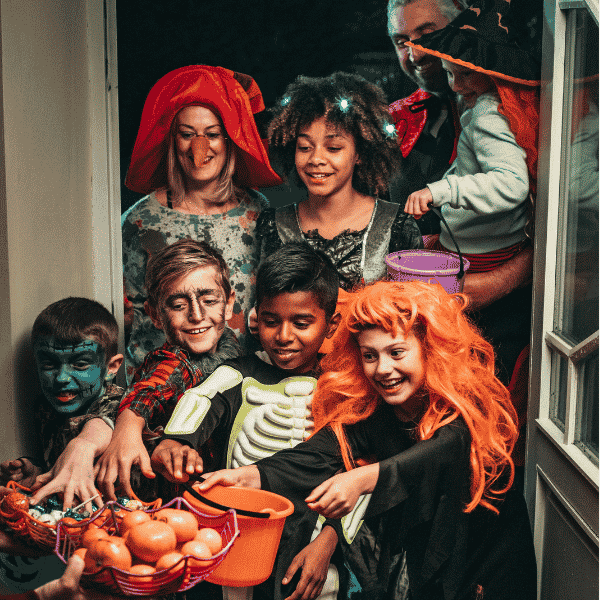 Children and adults in Halloween costumes crowd around a basket of candy at a doorway.