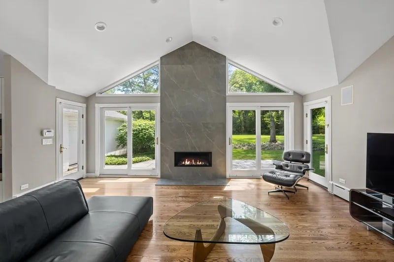 Living room with a tall stone fireplace, vaulted ceiling, and glass doors overlooking a green yard.