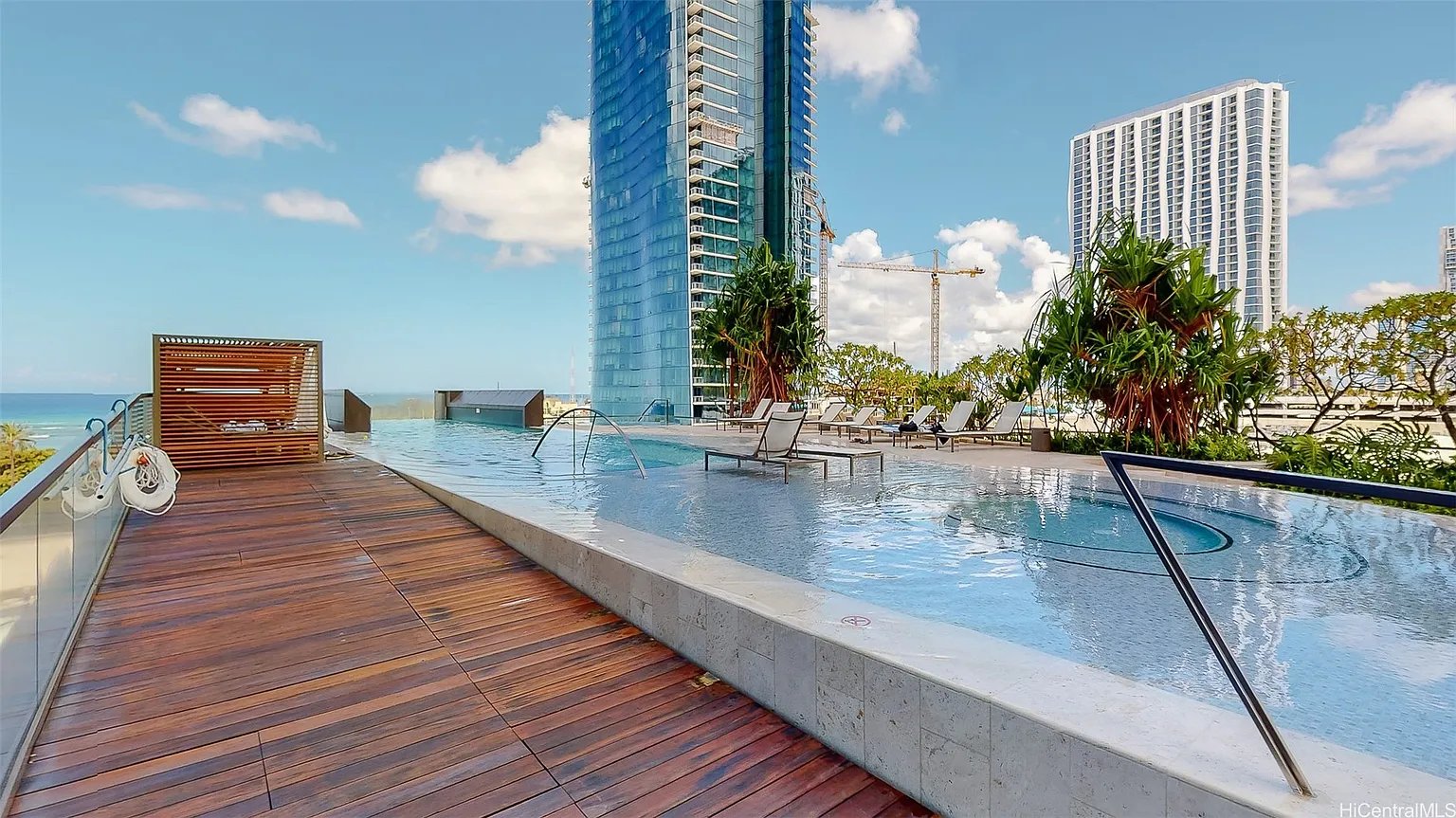 Rooftop pool beside a wooden deck, with lounge chairs, tropical plants, and ocean views.