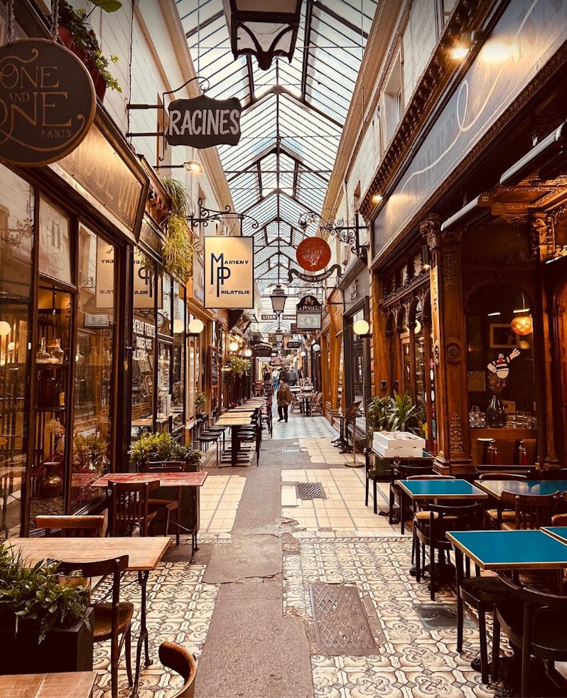 Parisian shopping arcade beneath a glass roof, lined with ornate shops and café tables.