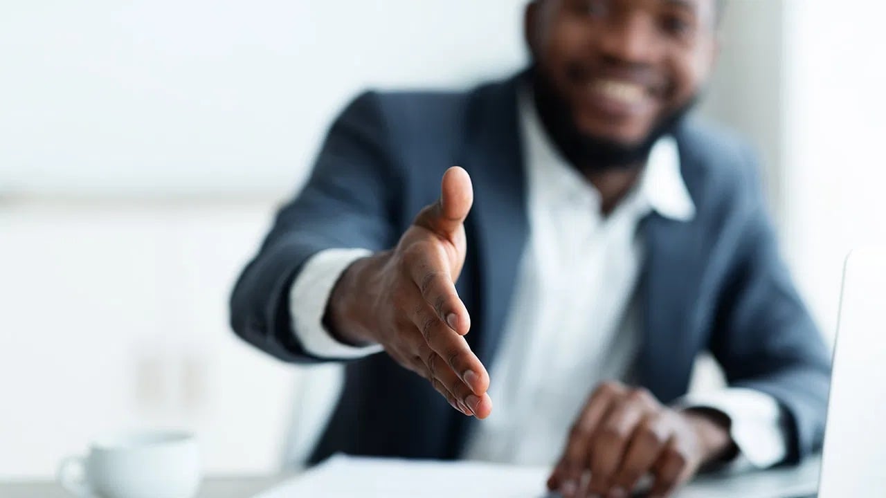 Smiling man in a dark suit reaching across a desk to offer a handshake.