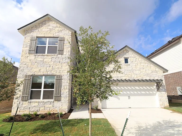 Two-story stone house with gray shutters, attached garage, and a young tree in the front yard.