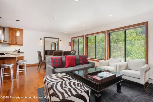 Living room with a gray sofa, white armchairs, patterned ottoman, and large windows overlooking trees.