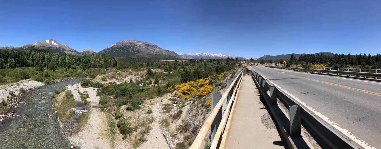 Rocky river and bridge with forested foothills and snow-dusted mountains beyond.