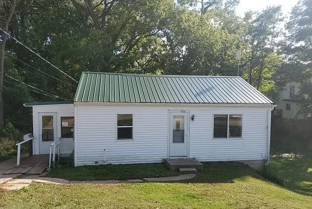 White single-story house with a green metal roof, front steps, and mature trees around it.