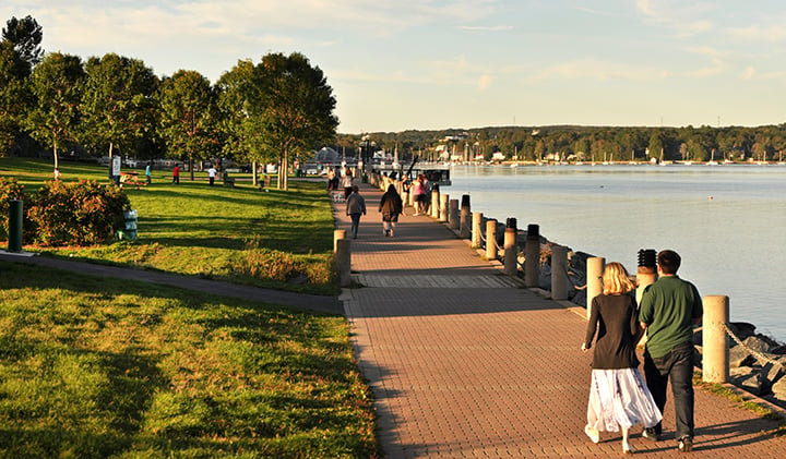 People stroll along a brick waterfront path beside a grassy park and calm water.