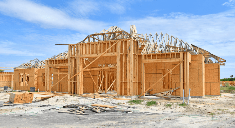 Wooden house frame under construction on a clear day. Exposed beams and trusses form the structure, conveying a sense of progress and development.