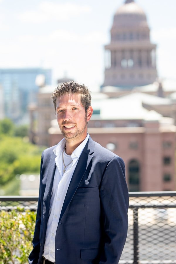 Man in a navy suit on a rooftop terrace, with the Texas State Capitol behind him.