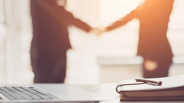 Laptop and paperwork on a desk as two people shake hands in the background.
