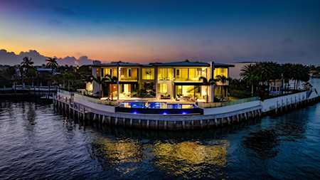Modern waterfront home glowing at dusk, with a pool and palm-lined dock.