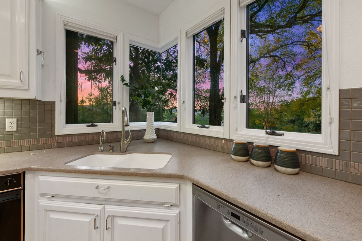 Kitchen sink area at 2131 Barton Hills Drive, Austin, TX 78704, with corner windows framing a sunset view through oak trees, granite counters and white cabinetry. Barton Hills neighborhood.