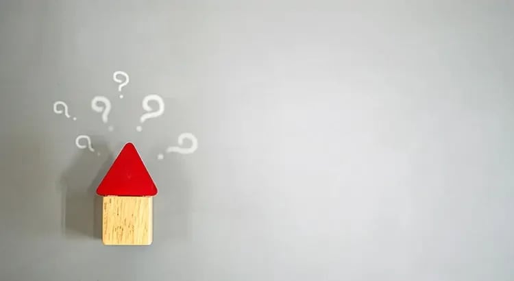 A small red-roofed house cutout with question marks around it, placed against a light gray background. It symbolizes questions or uncertainty related to housing or real estate.