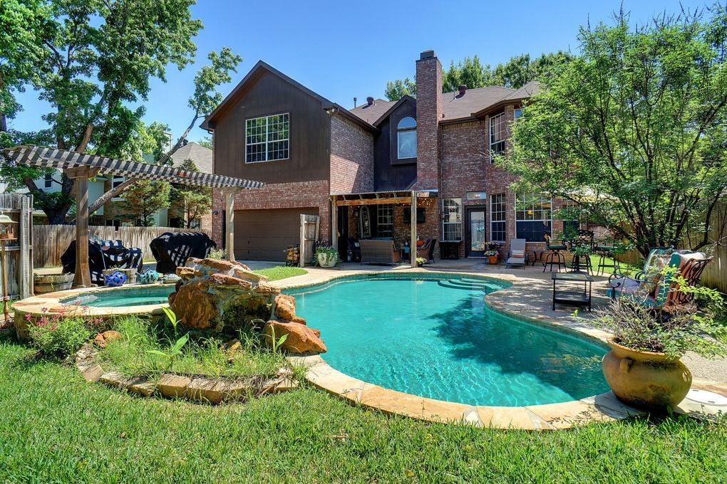 Two-story home behind a freeform pool, with a small spa and striped pergola.