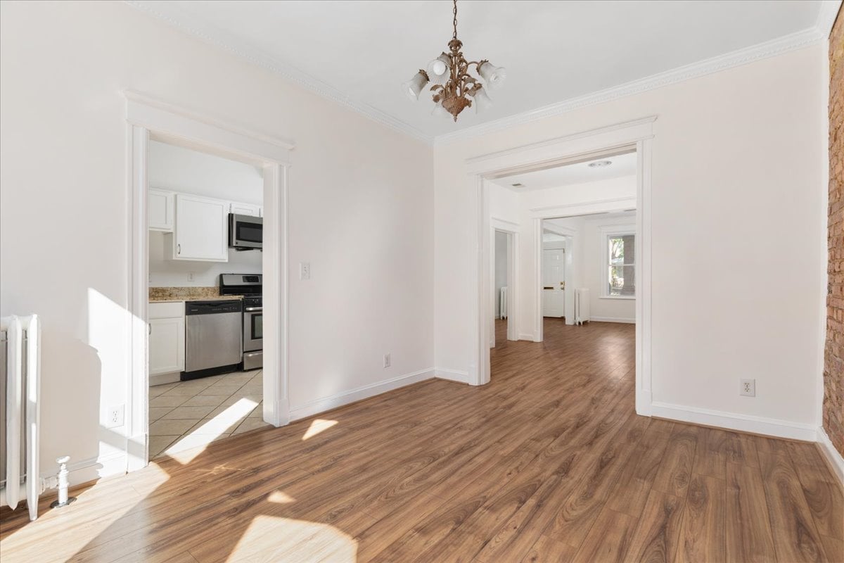Vacant room with hardwood floors, a chandelier, and doorways to the kitchen and adjoining room.
