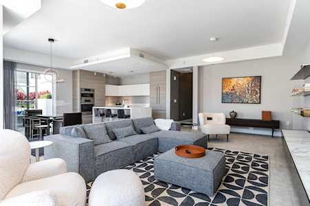 Open-plan living room with a gray sectional, patterned rug, and white accent chairs beside the kitchen.