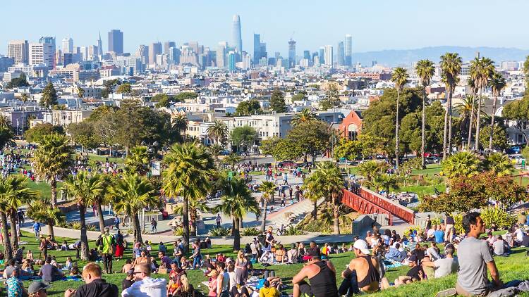 Panoramic view of Mission Dolores Park with people picnicking and enjoying the San Francisco skyline in the background.