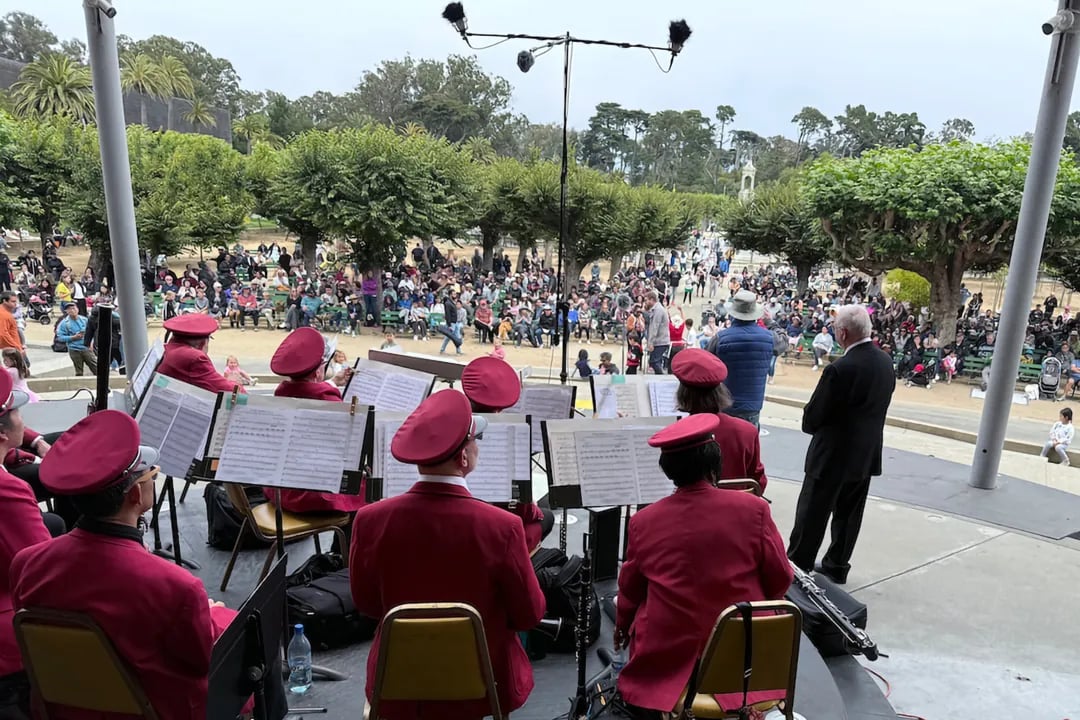 The Golden Gate Park Band in their signature red uniforms performing on stage for a large crowd gathered at the Music Concourse in San Francisco.