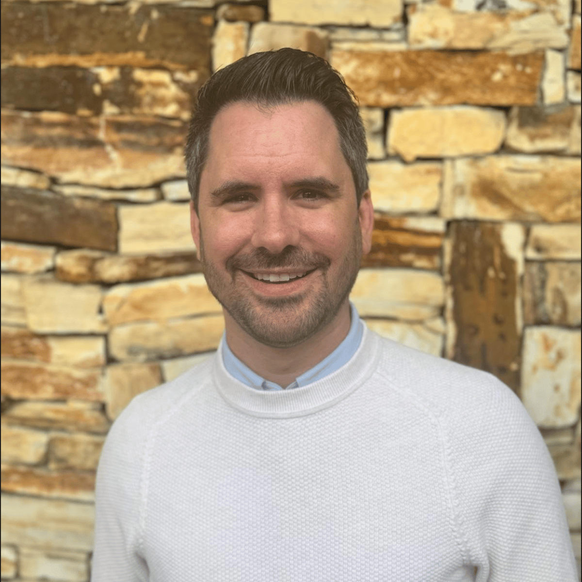 Andrew Miller smiling in a white sweater in front of a stone wall.