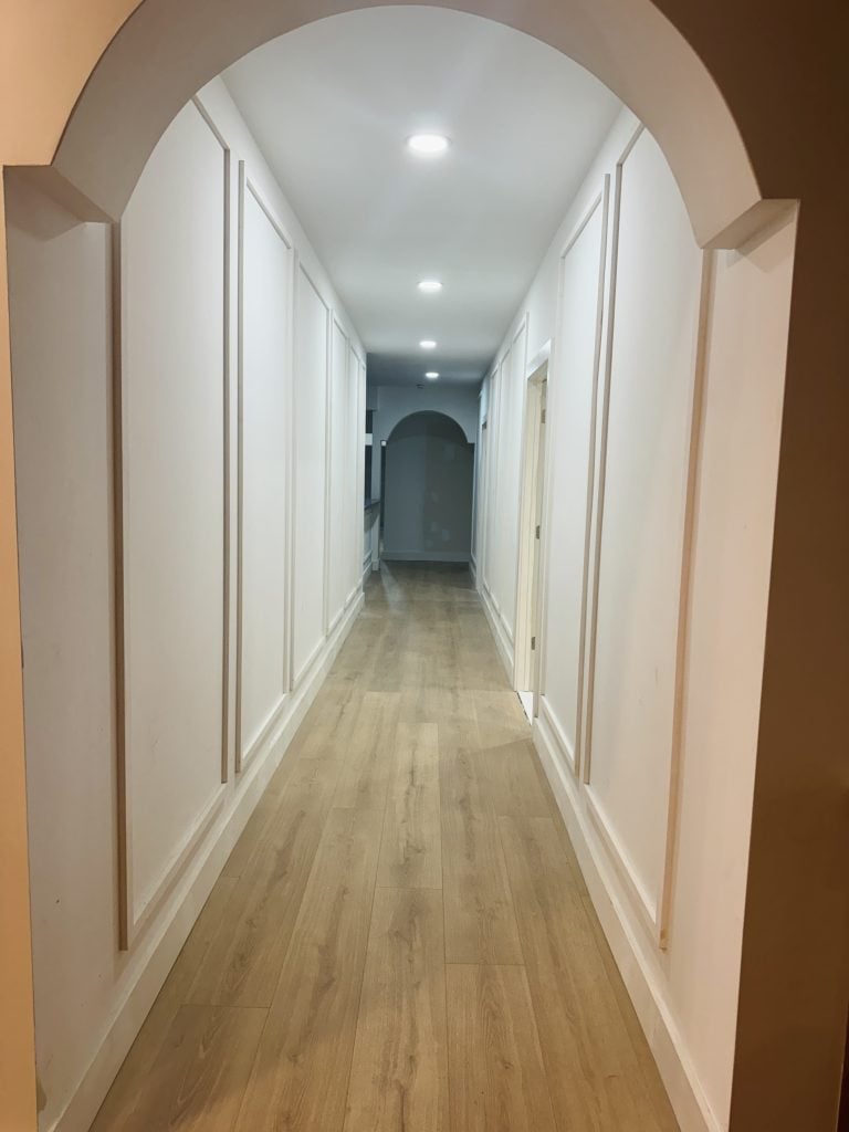 Renovated hallway with light wood flooring, white paneled walls, and recessed ceiling lights.
