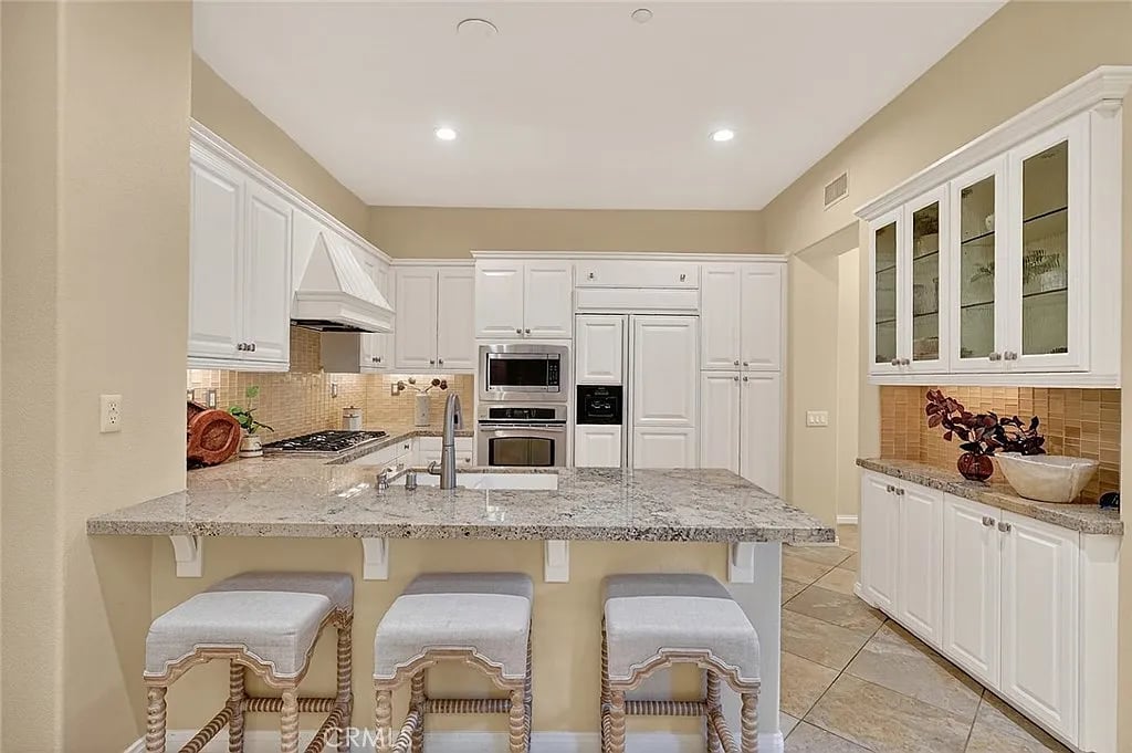 Kitchen with a granite island, three upholstered stools, white cabinetry, and built-in appliances.