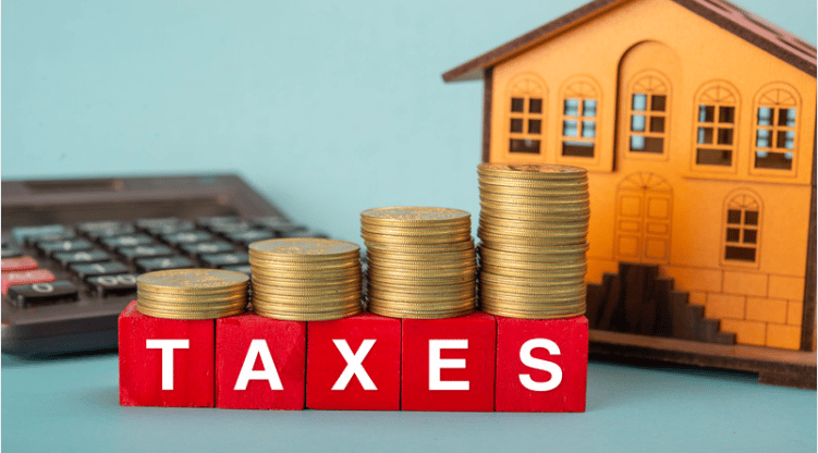 Stacks of coins on blocks spelling “TAXES,” with a model house and calculator behind them.