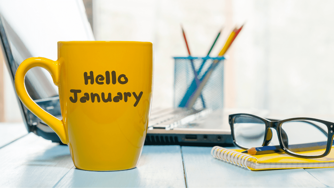 A yellow coffee mug with the text "Hello January" written on it. The mug is sitting on a desk or table, with a blurred background featuring a laptop, a pencil holder with colorful pencils, and a pair of glasses resting on a notebook.
