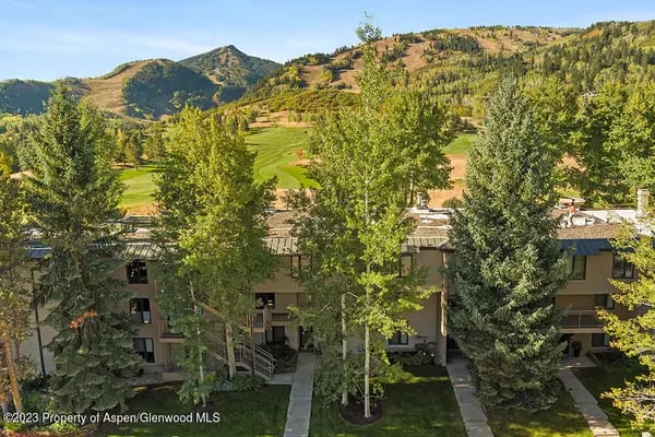 Multi-unit residential building among mature trees, with green mountain slopes behind it.