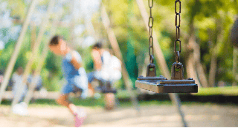 A playground swing in focus with two children playing in the blurred background.
