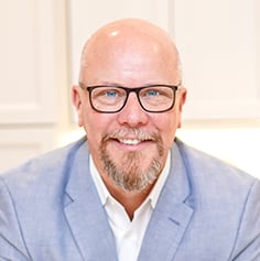 Bald man with glasses and a salt-and-pepper goatee wearing a light blue blazer.