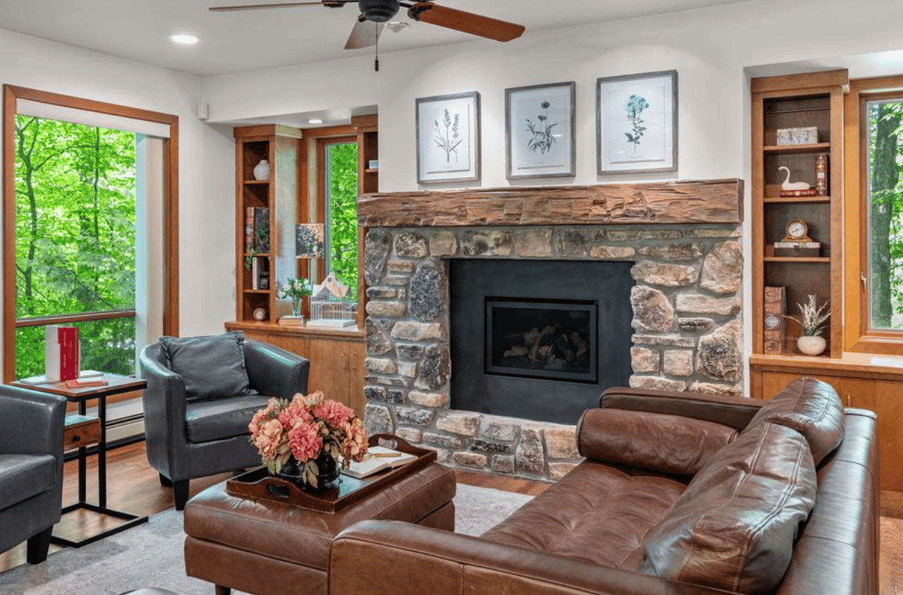 Living room with a stone fireplace, leather seating, and large windows overlooking trees.