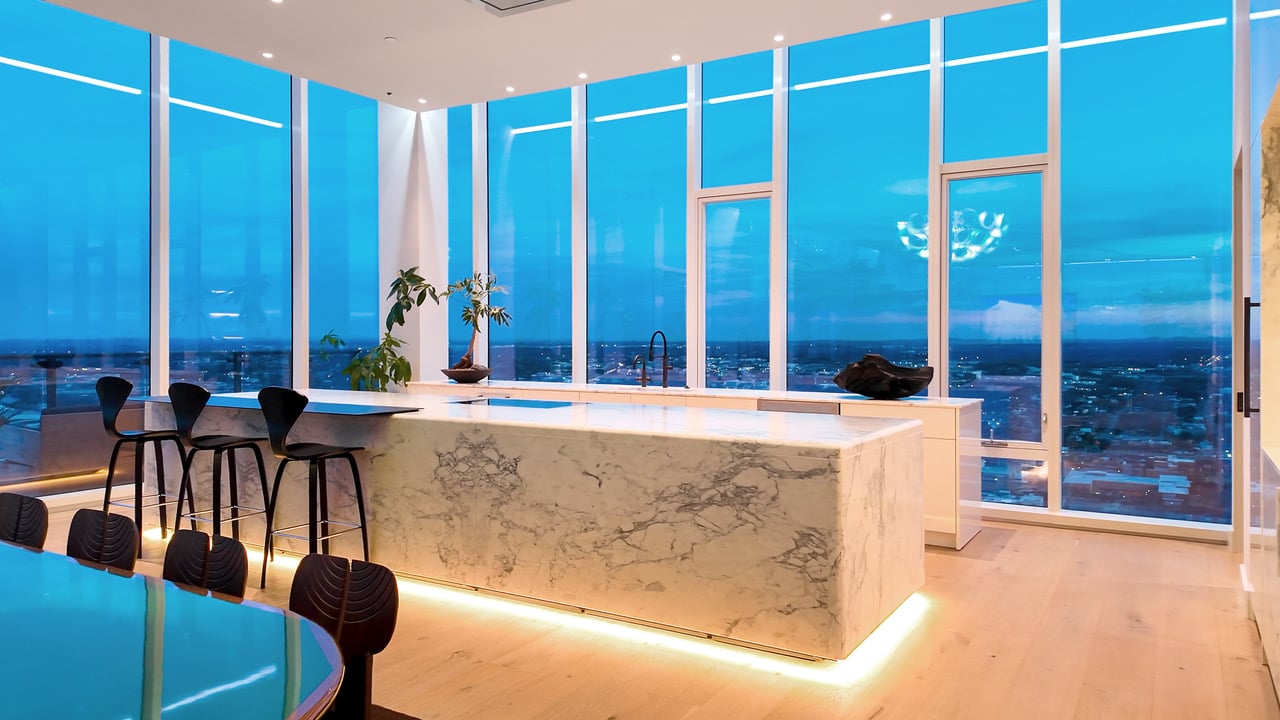 Modern kitchen with a marble island and floor-to-ceiling windows overlooking the city at dusk.