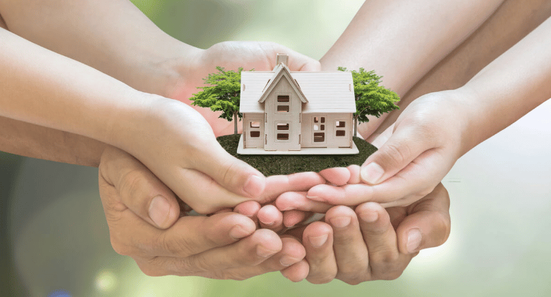 Several hands cradle a miniature house with small trees.