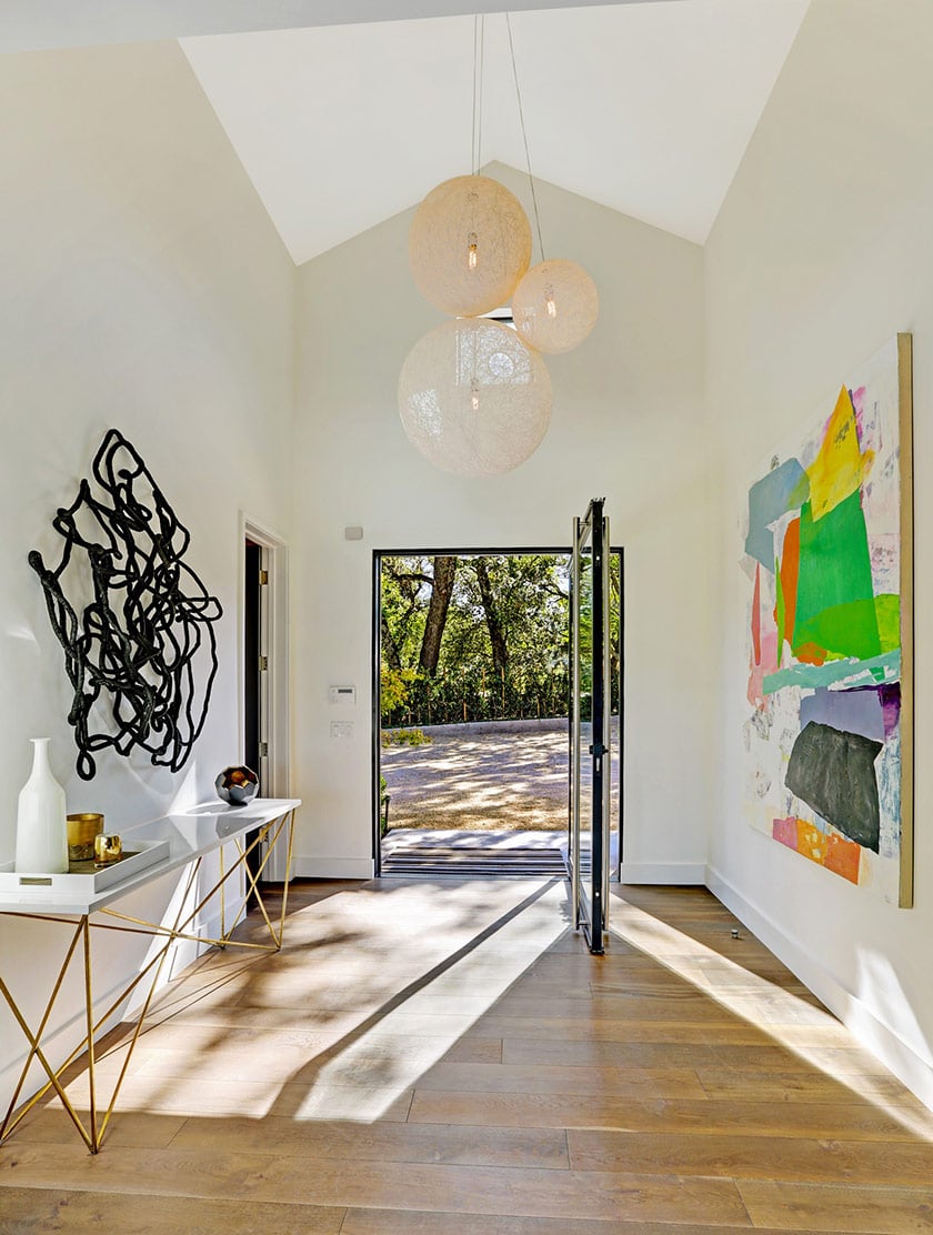 Bright entry foyer with globe pendants, abstract art, wood floors, and an open glass door to a terrace.