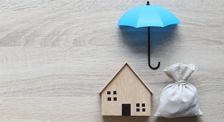 A small wooden house model with a blue umbrella over it and a bag of money beside it, representing insurance or financial protection for a home.