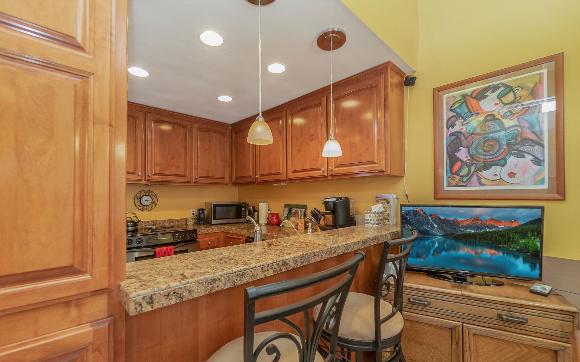 Kitchen with wood cabinets, granite breakfast bar, metal stools, and colorful framed artwork.