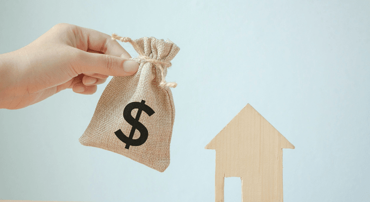 A hand holds a money bag beside a house model on a table, symbolizing financial investment in real estate.