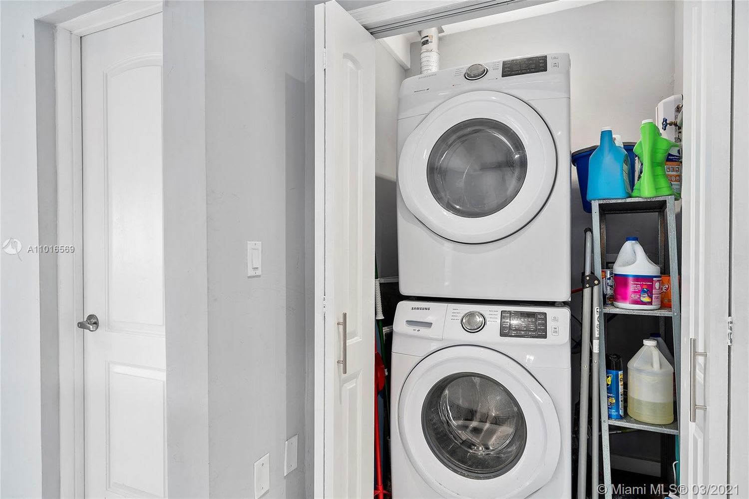 Laundry room with stacked white front-loading washer and dryer beside shelves of cleaning supplies.