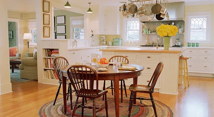 The kitchen has wooden chairs around a round table, bright yellow flowers in a vase, and pendant lights hanging from the ceiling. The overall ambiance is warm and inviting.