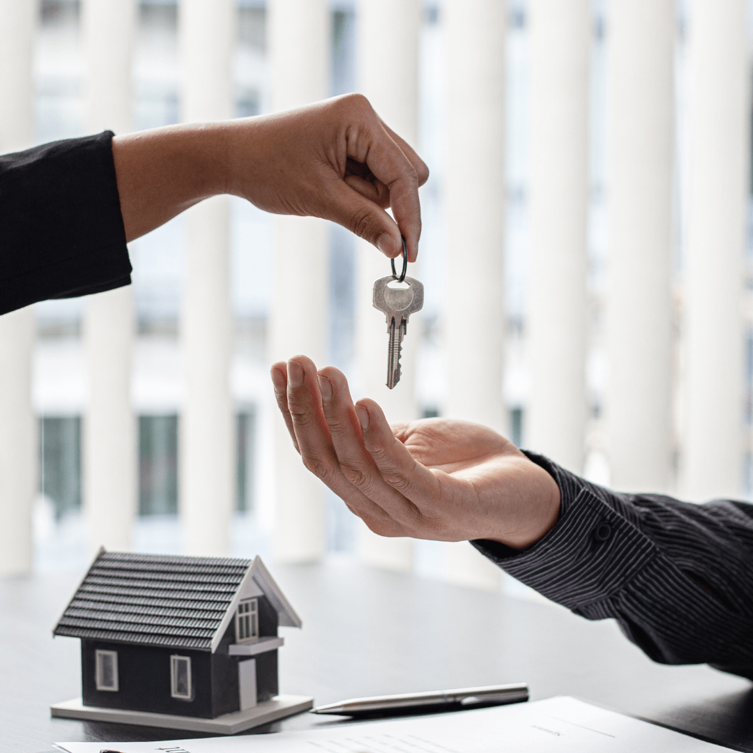 One person hands a key to another above a model house and real estate paperwork.