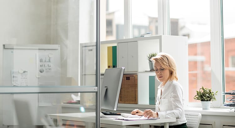 She is focused on papers in front of her, possibly reviewing or working on documents. The office is bright, with large windows letting in natural light. In the background, there are shelves with books, plants, and organizational items, adding to the professional but minimalist atmosphere. The overall setting conveys a productive and focused work environment.