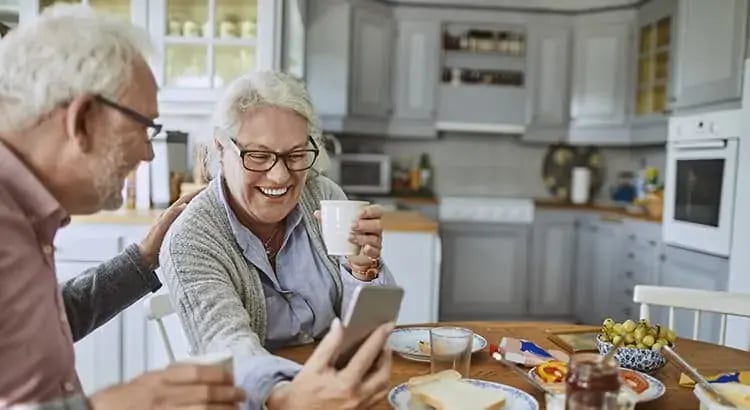 An elderly couple enjoying a pleasant moment together in their kitchen. They are sitting at a kitchen table, smiling and laughing as they look at something on a smartphone that the woman is holding.